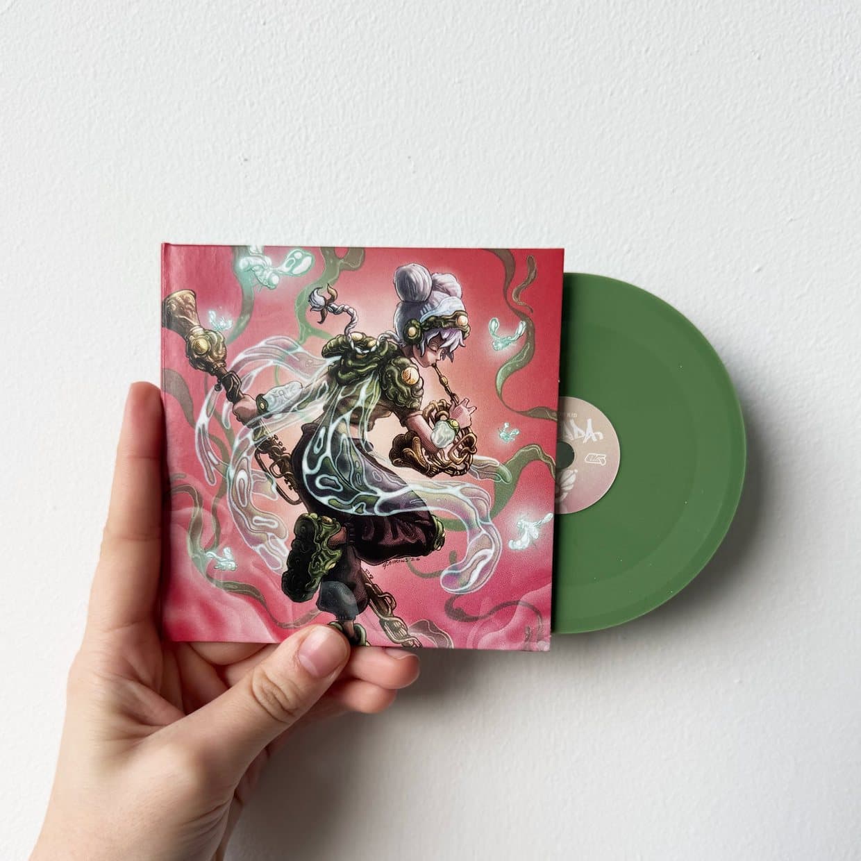 A hand holding a pink illustrated record sleeve with a green vinyl record sliding out of it, against a pale wall