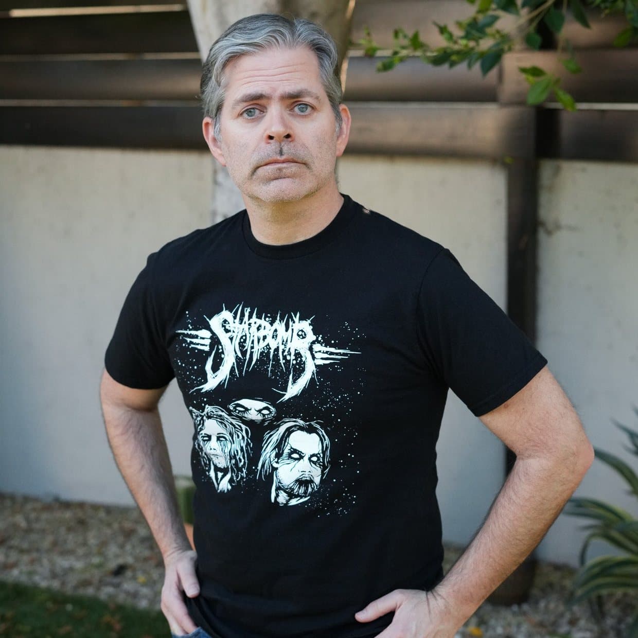 A man standing outdoors in front of a hedge, wearing the black Starbomb tee with its white band logo and two faces across the chest