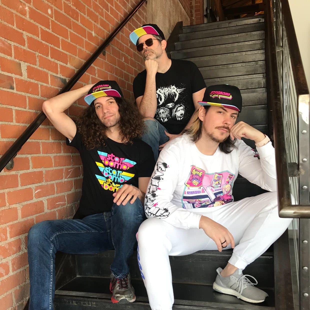 Three people sitting on an iron staircase against a brick wall, wearing Starbomb caps and printed tees
